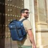 Mann mit dunkelblauem Cabin Max Metz Rucksack auf dem Rücken, ideal für Handgepäck bei Wizz Air und Eurowings, vor historischem Hintergrund.