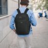 Junger Mann mit einem schwarzen ECOHUB 20L Handgepäck Rucksack, der urbanes Umfeld genießt. Der Rucksack ist geräumig und sicher, ideal für Reisen. Er trägt ein hellblaues Hemd und beige Hosen.