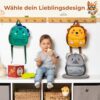 Fröhlicher Junge mit einem grauen T-Shirt und Jeans, der auf einer Bank sitzt und ein Stück Essen hält, vor einer Auswahl von bunten Kinder-Rucksäcken in Tierdesigns, darunter ein Dinosaurier, ein Löwe und ein Koala.