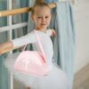 Mädchen im Ballettanz-Outfit mit rosa Rucksack, der eine Schleife und ein Ballettmotiv zeigt, in einem Tanzstudio mit natürlichem Licht.