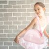 Fröhliches Mädchen in einem Ballettanzug mit rosa Umhängetasche, die einen dekorativen Ballettschuh zeigt, vor einer modernen Ziegelwand.