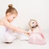 Kleines Mädchen in einem Ballett-Outfit untersucht eine rosa Halbmond-Tasche, umgeben von einem Plüschbären, vermittelt Unschuld und Freude am Spielen.